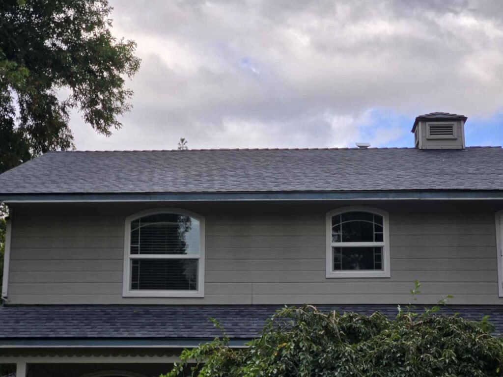 Two-story residential home with newly installed asphalt shingle roof, gray lap siding, and white-framed arched windows under an overcast sky.