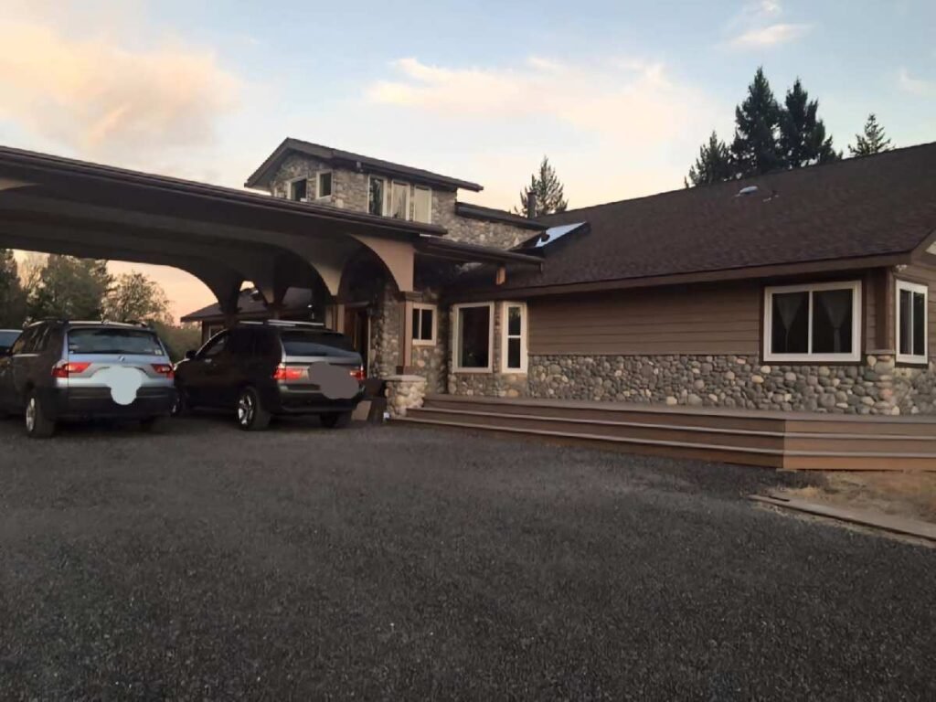 Exterior of a custom home with stone and wood siding, covered carport with two parked vehicles, dark shingle roof, gravel driveway, and trees in the background at sunset.