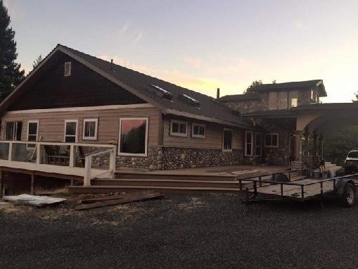 Residential home exterior under construction with new siding and large windows installed at dusk, showing an unfinished deck area and gravel ground.